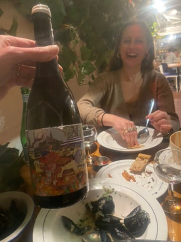 Person holding a wine bottle at a dinner table with mussel shells, while a woman smiles in the background
