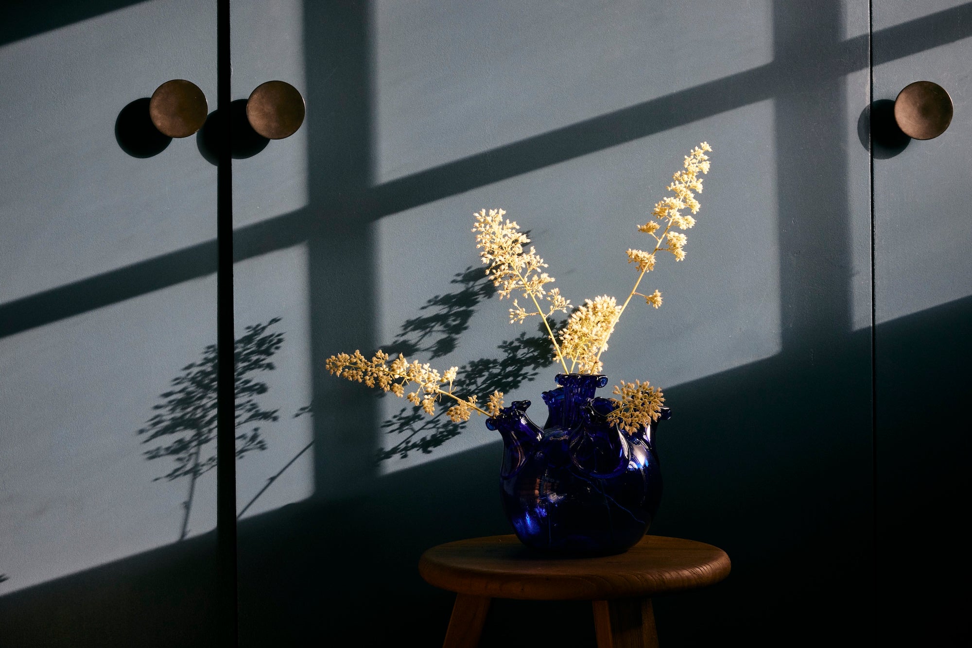 The Lola Tulip Vase in dark blue, filled with dried yellow flowers, rests on a wooden stool and casts shadows on a gray wall lit by sunlight streaming through a window.