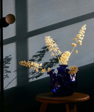 The Lola Tulip Vase in dark blue, filled with dried yellow flowers, rests on a wooden stool and casts shadows on a gray wall lit by sunlight streaming through a window.