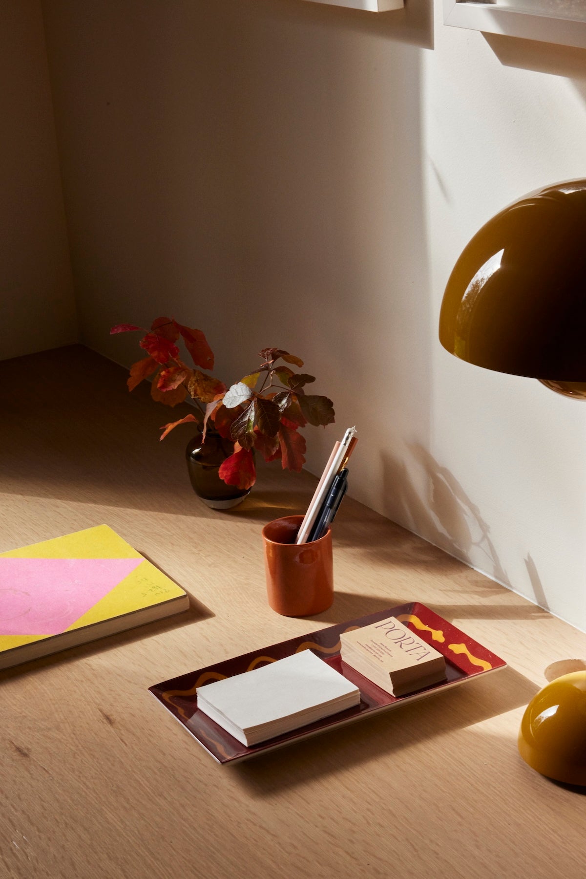 A wooden desk in sunlight, featuring a yellow and pink notebook, the Hand Painted Double Serpent Tray holding paper and cards, a cup with pens, a small potted plant, and a brown lamp.