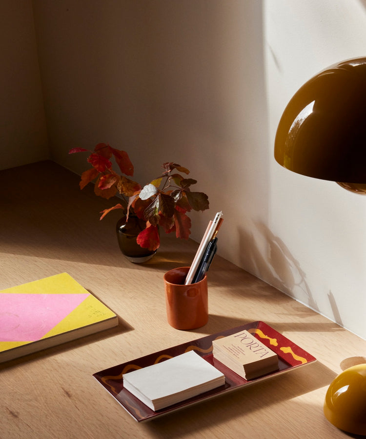 A wooden desk in sunlight, featuring a yellow and pink notebook, the Hand Painted Double Serpent Tray holding paper and cards, a cup with pens, a small potted plant, and a brown lamp.
