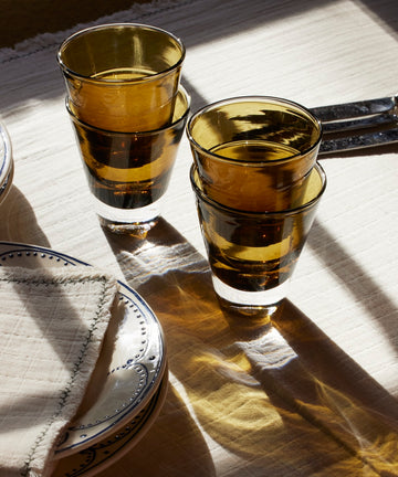 Two stacks of Gem Glasses, Set of 2, in amber conical shapes, sit on a sunlit tablecloth beside two plates, a folded napkin, and utensils—evoking the relaxed charm of the South of France.