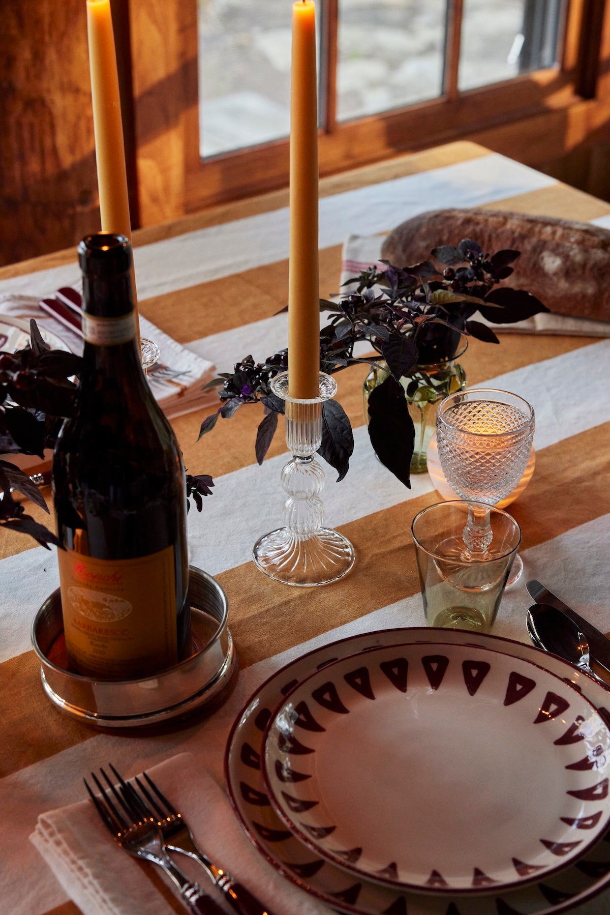 A dining table set near a window features patterned plates, silverware, a wine bottle, candles, glassware, and small floral arrangements atop the artisanal Riga Tablecloth.