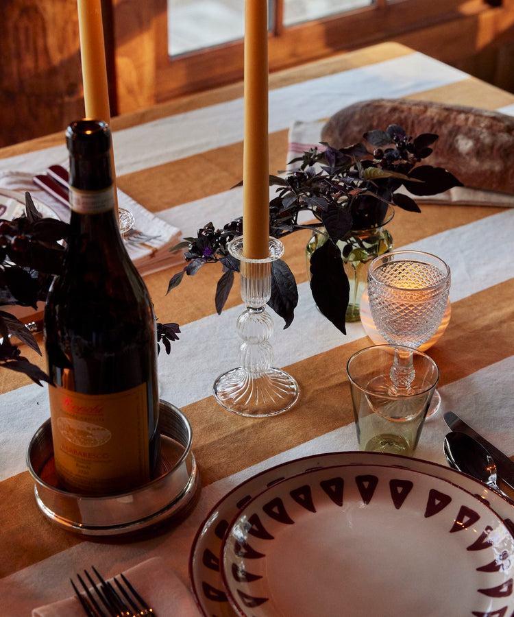A dining table set near a window features patterned plates, silverware, a wine bottle, candles, glassware, and small floral arrangements atop the artisanal Riga Tablecloth.