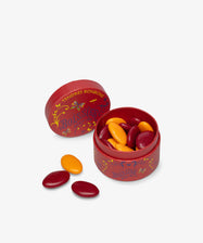 A round red Boissier box holds Multicolored Chocolate Gems, with its lid beside it and a few gems scattered outside.