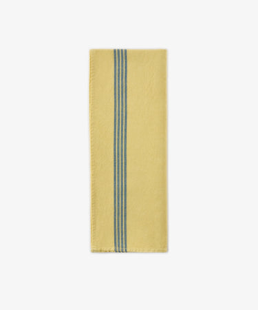 A Métis Tea Towel, folded to showcase its yellow fabric accented by two parallel blue woven stripes running vertically near one edge—a design evocative of classic French kitchen linens—is displayed on a white background.