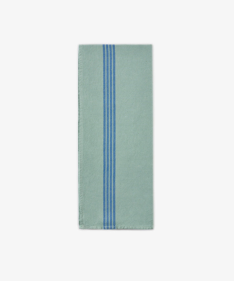 The Métis Tea Towel, a folded light green cloth featuring three vertical blue stripes near the center, is displayed on a plain white background and evokes the look of French kitchen linens and classic vintage striped tea towels.