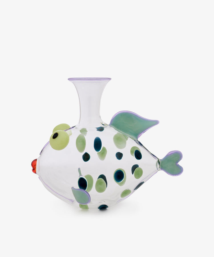 The Dotfish Decanter—crafted from clear glass and accented with green and blue spots, green fins, a red mouth, and raised round eyes—is inspired by Murano glass techniques. This whimsical fish-shaped vase is shown against a plain white background and evokes the playful design of Massimo Lunardon’s iconic Dotfish Decanter.