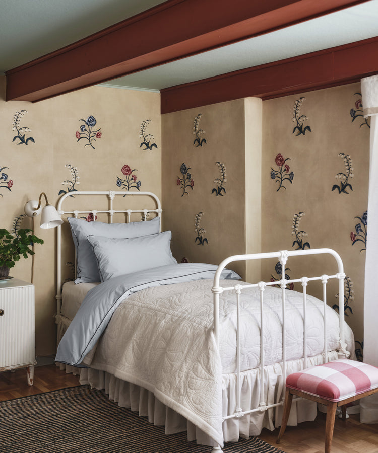 A Dawn Twin Bedspread in light blue covers a neatly made white metal bed set against a beige floral-patterned wall in a cozy bedroom with exposed red beams.