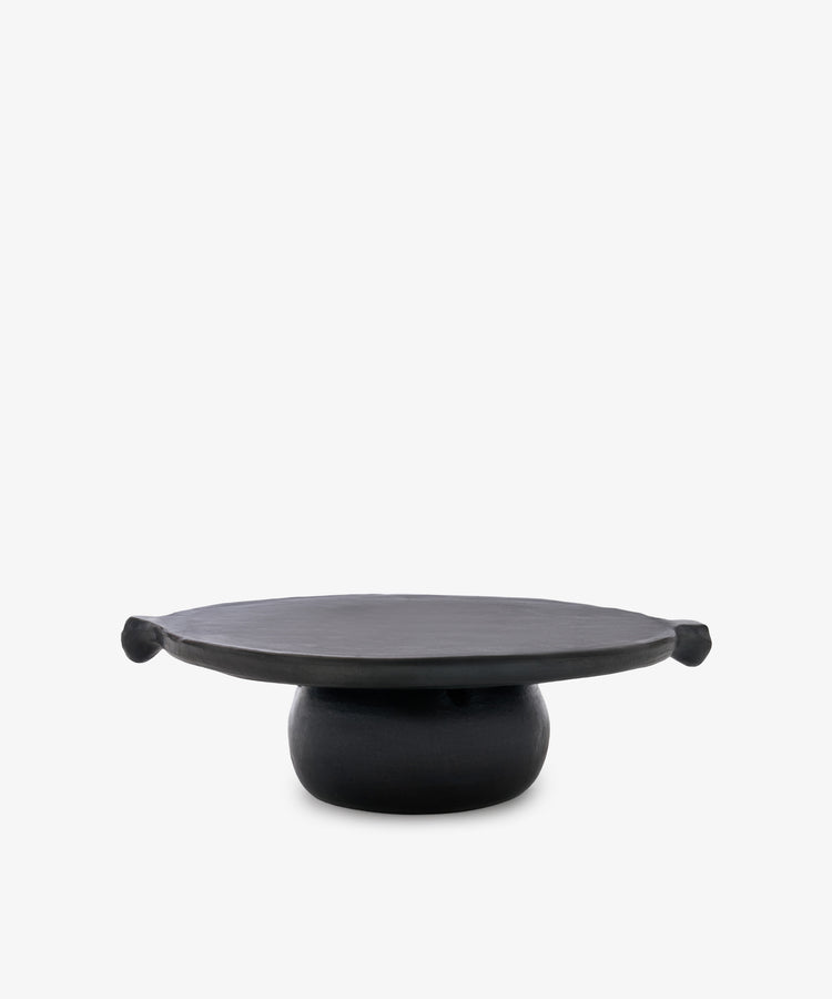 The Chamba Fruit Bowl is a black, round Colombian pottery pedestal plate with a wide flat top and two knob-like handles, beautifully handmade.