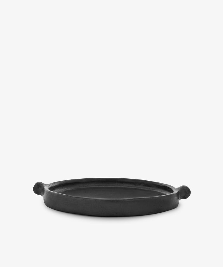 The Chamba Plate is a round, flat black clay plate with two small handles, handmade in Colombia and shown on a plain white background.