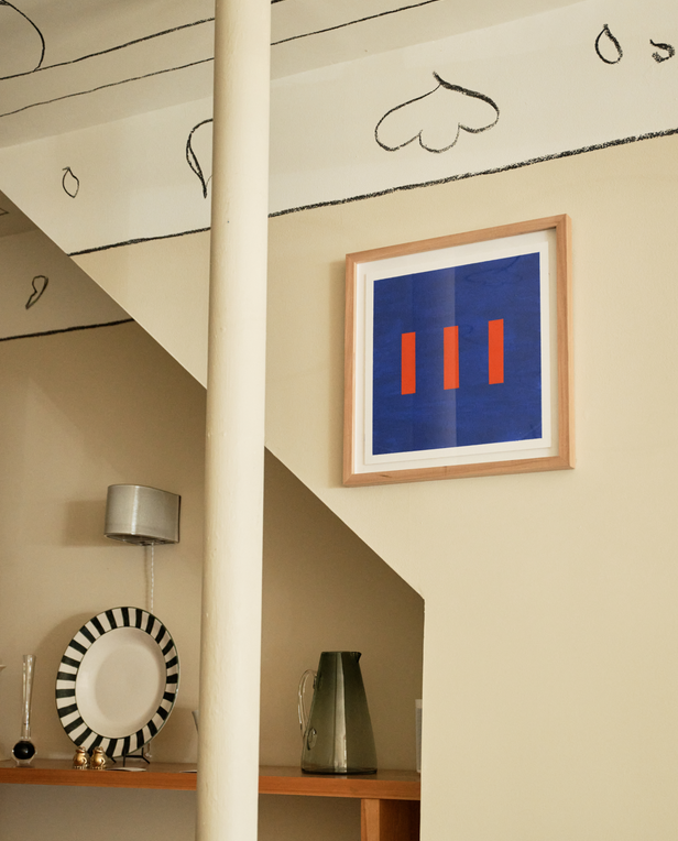 Framed abstract artwork with three red bars on a blue background hanging on a beige wall