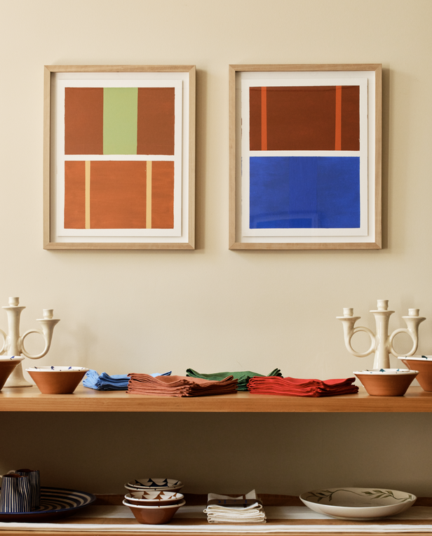 Two framed abstract artworks hang above a shelf with folded napkins, bowls, and candlesticks