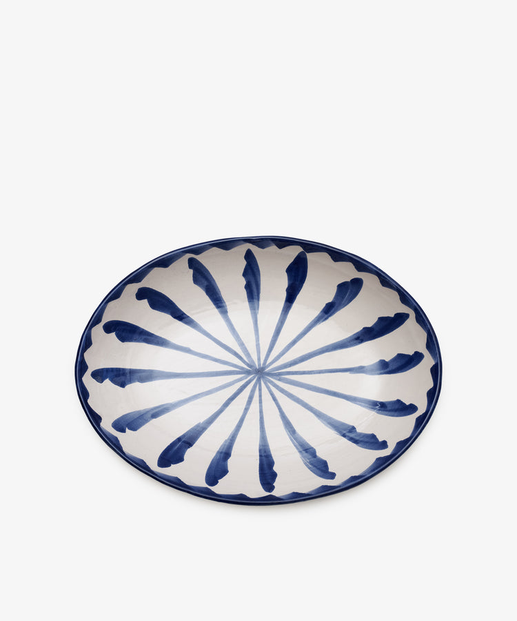 The Rayas Oval Serving Platter is a handmade ceramic piece featuring a blue and white radial feather-like pattern on the interior, highlighting its unique artistry.