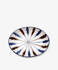 The Rayas Oval Serving Platter features alternating blue and brown brushstroke patterns radiating from the center on a white ceramic background, offering vibrant color and handmade appeal.