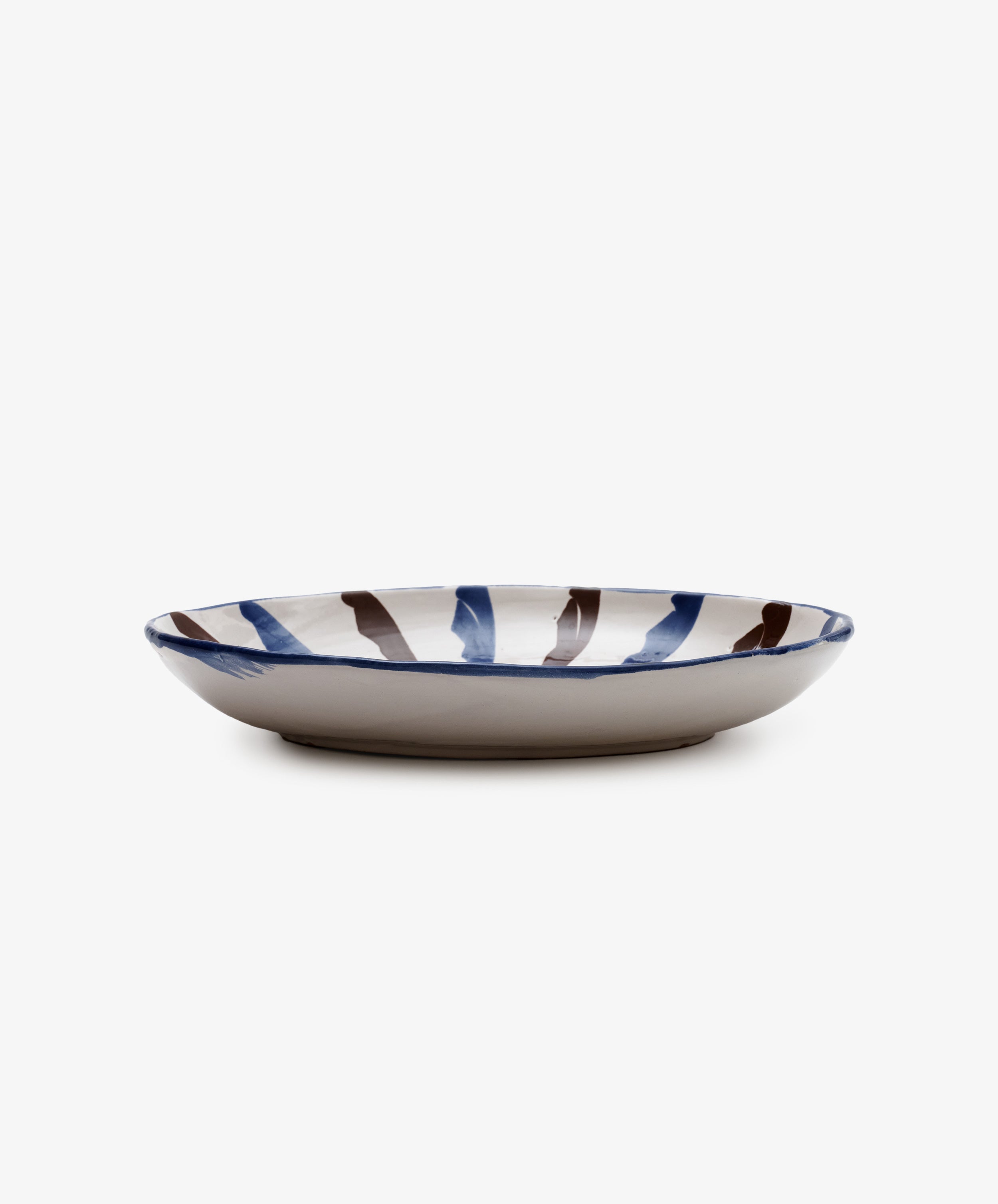 The Rayas Oval Serving Platter is a shallow, handmade ceramic plate featuring a white base with vibrant hand-painted blue and brown vertical stripes.