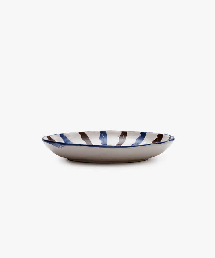 The Rayas Oval Serving Platter is a shallow, handmade ceramic plate with a beige base and blue and brown brushstrokes around the rim, displayed against a plain white background.