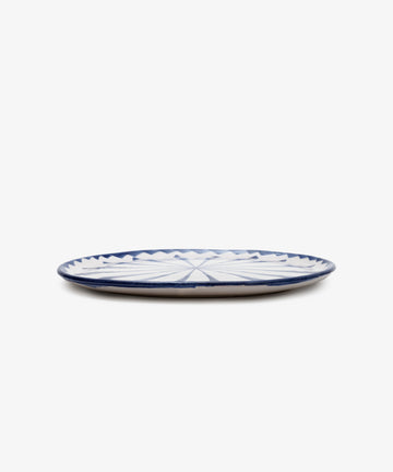 The Rayas Serving Plate is a flat, round handmade ceramic plate featuring a vibrant blue and white geometric pattern, displayed on a white background.