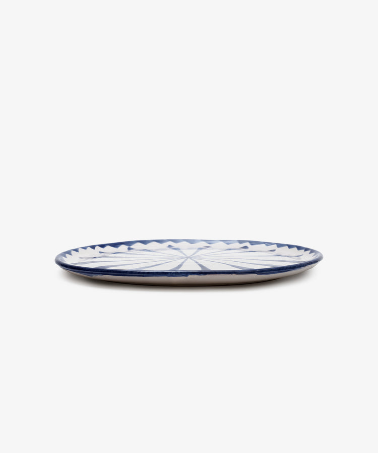 The Rayas Serving Plate is a flat, round handmade ceramic plate featuring a vibrant blue and white geometric pattern, displayed on a white background.