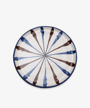 The Rayas Serving Plate is a round, hand-painted ceramic plate with a blue rim and alternating blue and brown radial stripes on a white background, bringing artistic flair to your table.