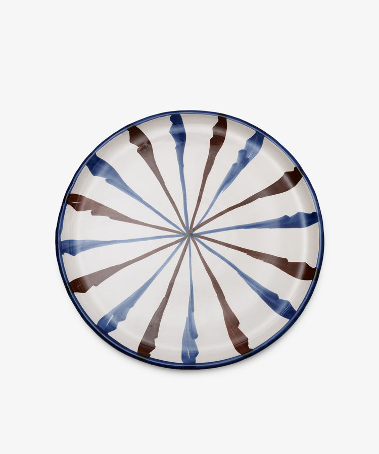 The Rayas Serving Plate is a round, hand-painted ceramic plate with a blue rim and alternating blue and brown radial stripes on a white background, bringing artistic flair to your table.