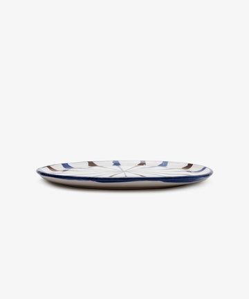 The Rayas Serving Plate is a low, round handmade plate with a white base and abstract blue and brown line patterns, shown against a plain white background.
