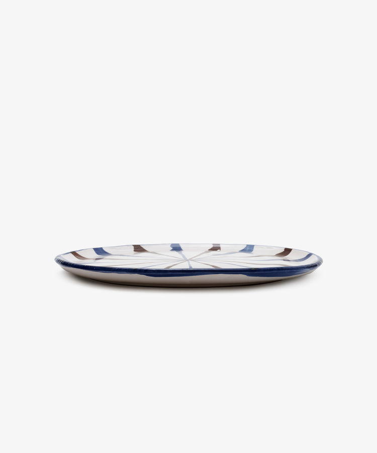 The Rayas Serving Plate is a low, round handmade plate with a white base and abstract blue and brown line patterns, shown against a plain white background.