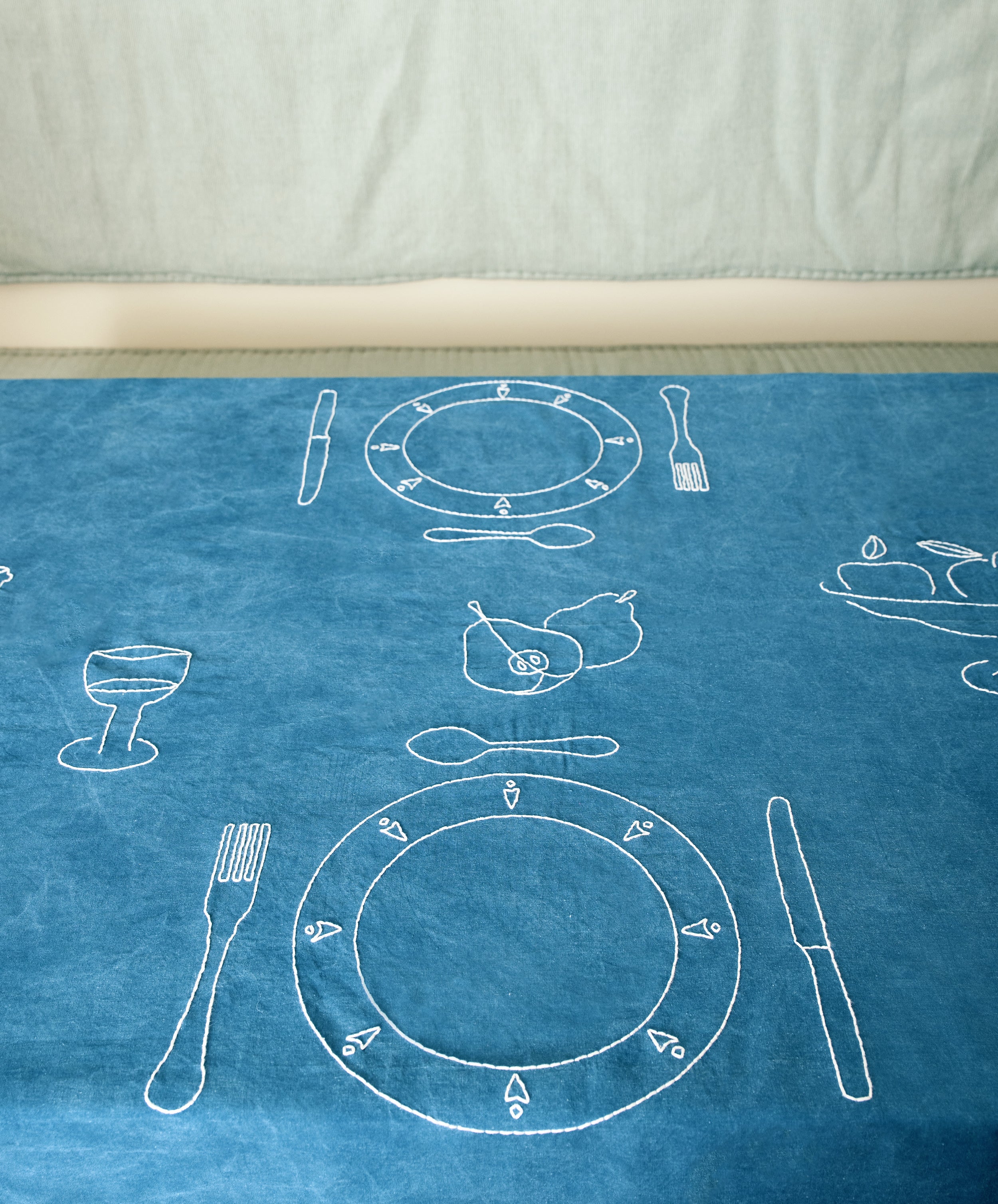 Dîner d'Eté custom tablecloth in indigo linen spread on table, embroidered with white thread place setting motifs.