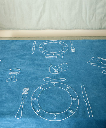 Dîner d'Eté custom tablecloth in indigo linen spread on table, embroidered with white thread place setting motifs.