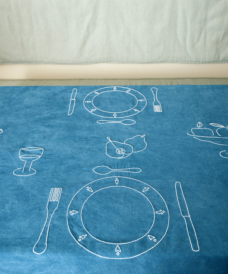 Dîner d'Eté custom tablecloth in indigo linen spread on table, embroidered with white thread place setting motifs.