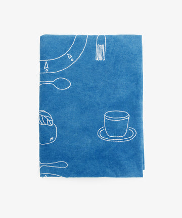 The Dejeuner d'Eta | Custom Tablecloth is a folded, blue hand-dyed cloth featuring white line drawings of a cup, saucer, fork, spoon, and food item, displayed on a plain white background.