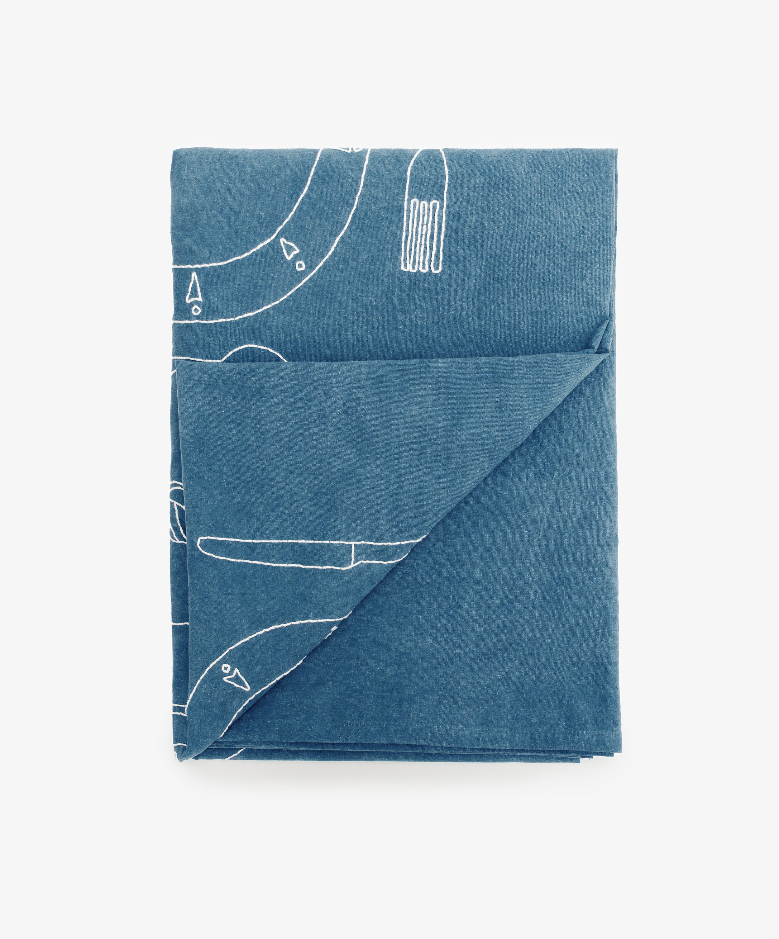 The Dejeuner d'Eta | Custom Tablecloth is a folded, hand-dyed blue tablecloth featuring white line drawings of a fork, knife, and abstract shapes.