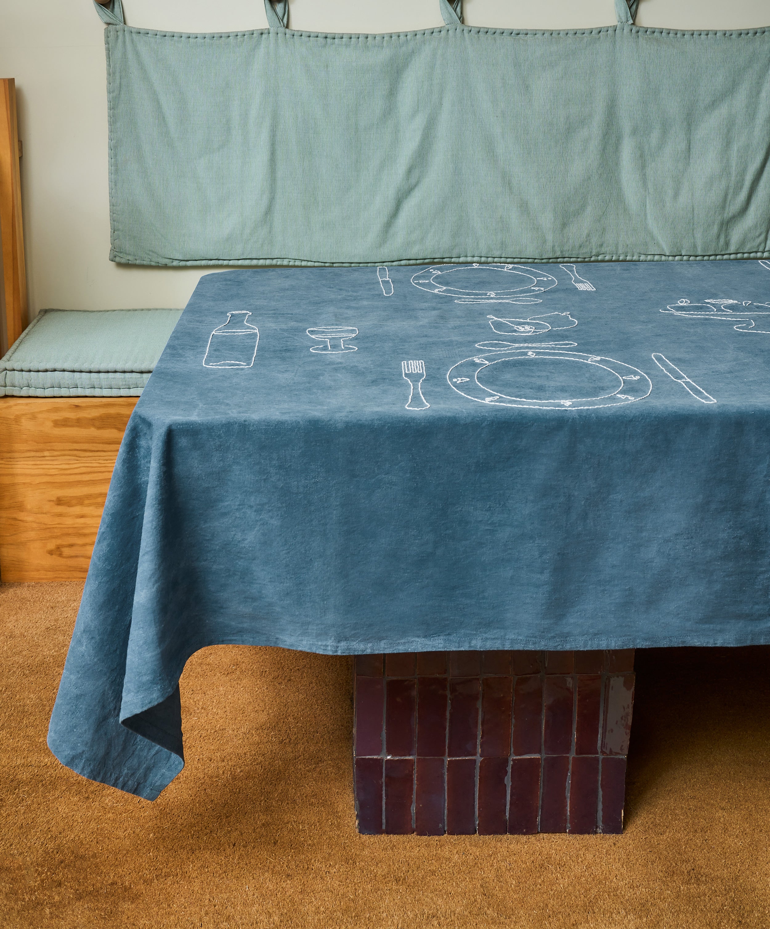 The Dejeuner d'Eta | Custom Tablecloth, featuring white line drawings of tableware on vintage linen, covers a rectangular table with a tiled base beside a wooden bench with blue-gray cushions.