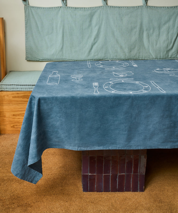 The Dejeuner d'Eta | Custom Tablecloth, featuring white line drawings of tableware on vintage linen, covers a rectangular table with a tiled base beside a wooden bench with blue-gray cushions.