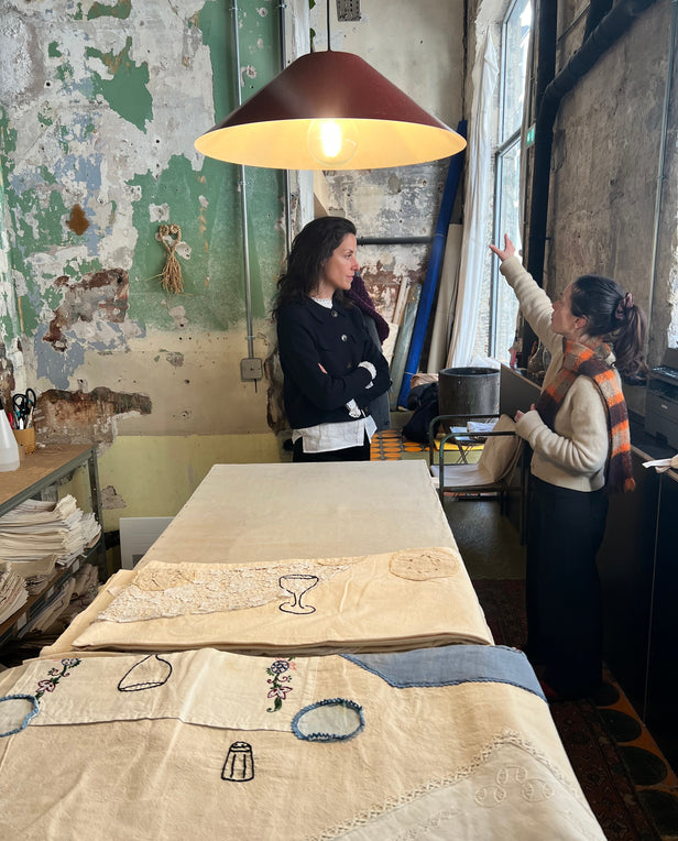 Two women talking in a rustic workshop with embroidered fabrics on a table