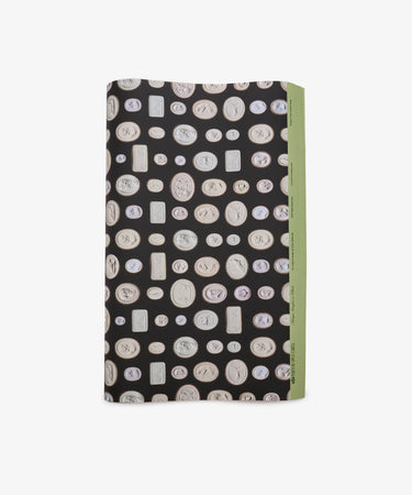 A rectangular object wrapped in Intaglio Gift Wrap, featuring black paper decorated with white coin-like motifs and accented by a green edge on the right, reminiscent of the luxurious packages used for Grand Tour gifts.