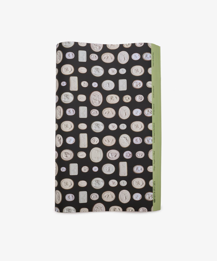 A rectangular object wrapped in Intaglio Gift Wrap, featuring black paper decorated with white coin-like motifs and accented by a green edge on the right, reminiscent of the luxurious packages used for Grand Tour gifts.