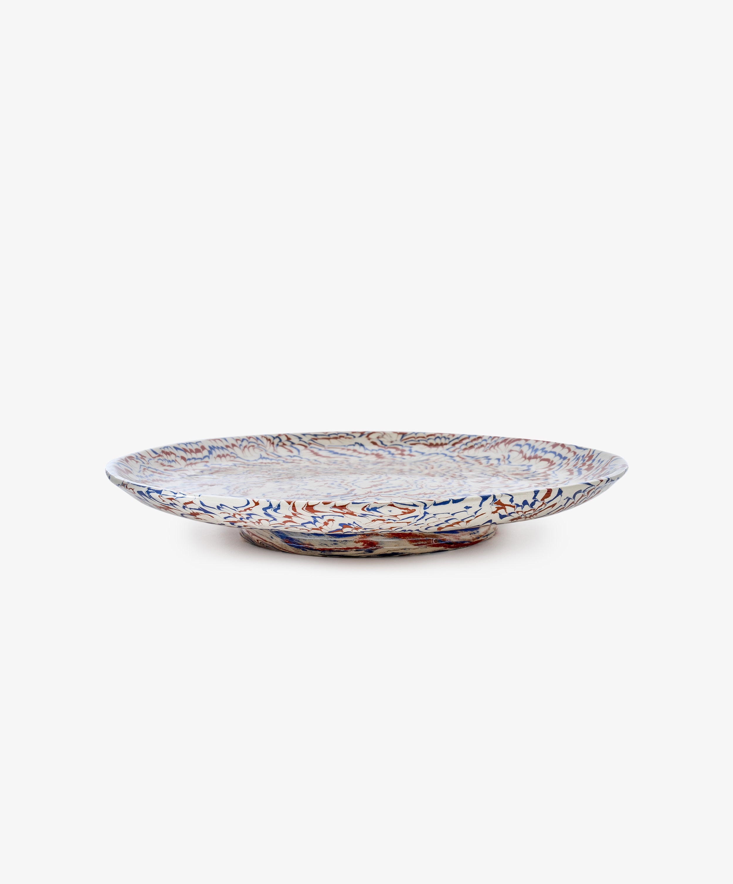 The Celine Serving Platter, a shallow ceramic plate reminiscent of a handmade serving dish, features intricate blue, red, and beige patterns on a white background and is shown from the side against a plain white backdrop.
