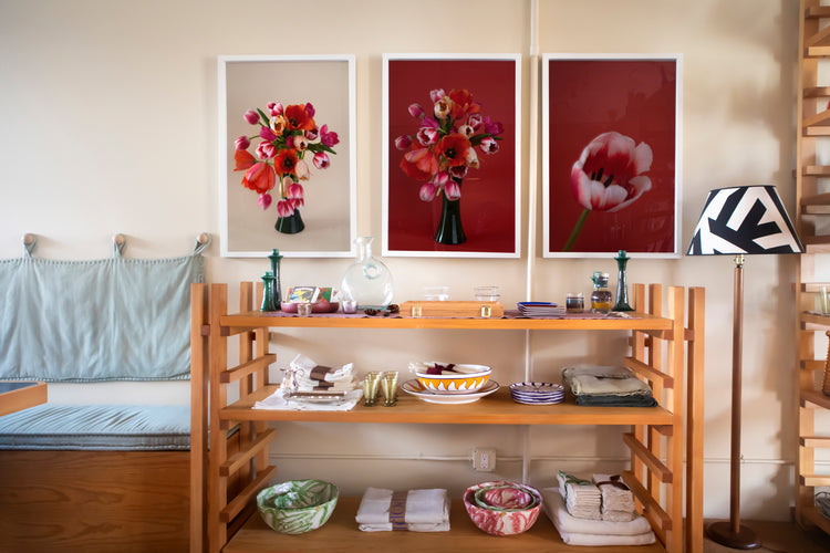 Wooden shelving unit with bowls, plates, and folded linens, set against a wall with three framed floral prints in red tones.