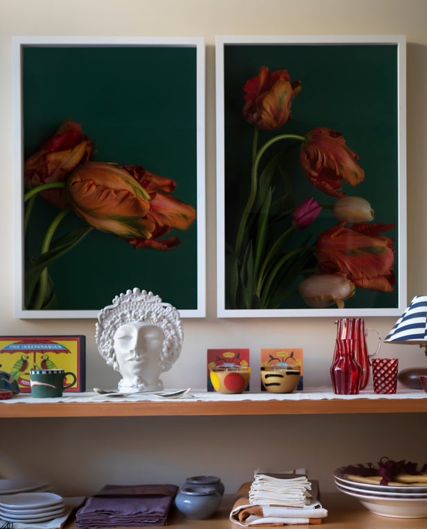 Wooden shelving unit with bowls, plates, and folded linens, set against a wall with three framed floral prints in red tones