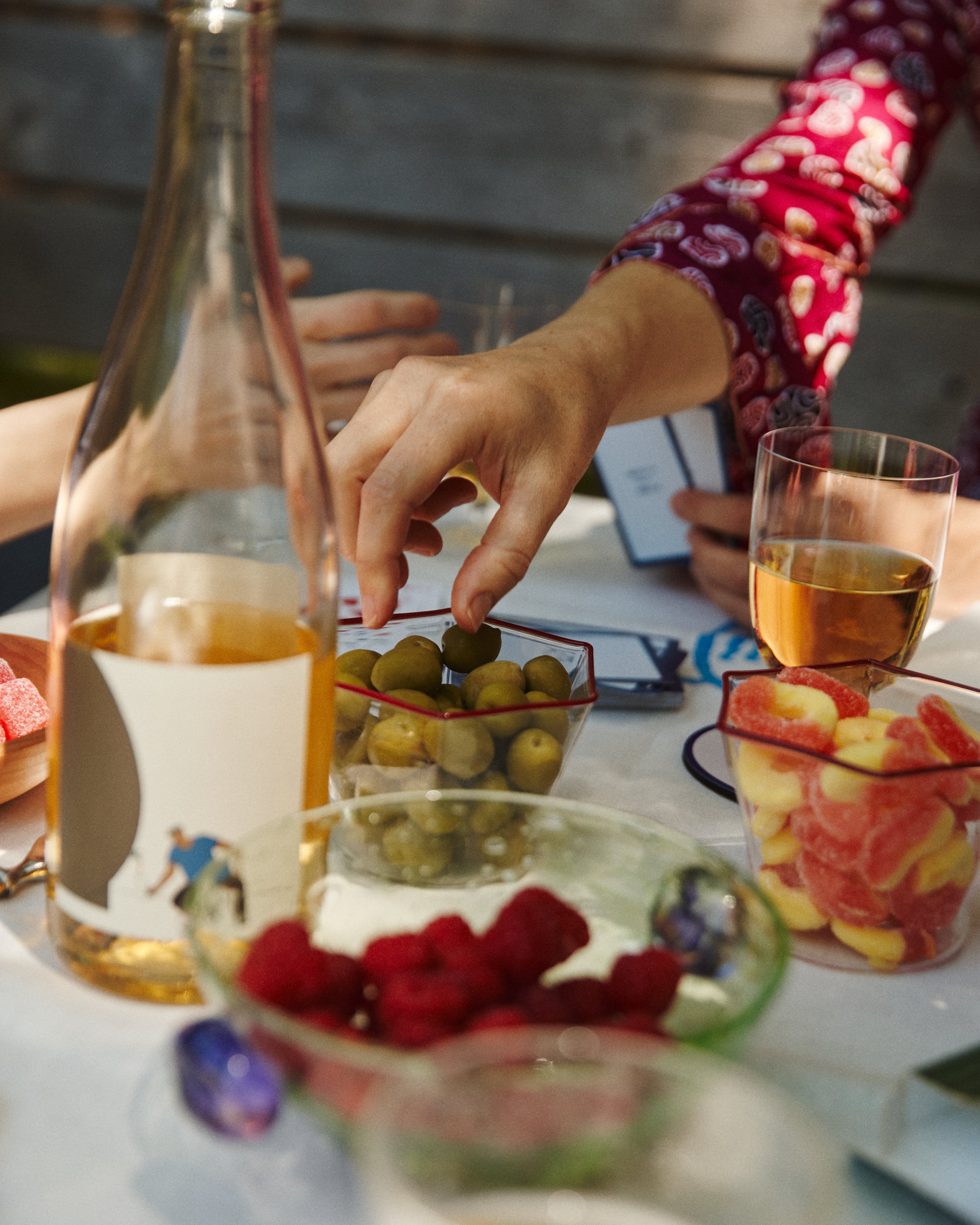 A hand reaches for green olives in a Gustavo Snack Bowl on an outdoor table set for aperitivo hour with wine, raspberries, gummies, elegant tableware, and glasses, while another person holds cards in the background.