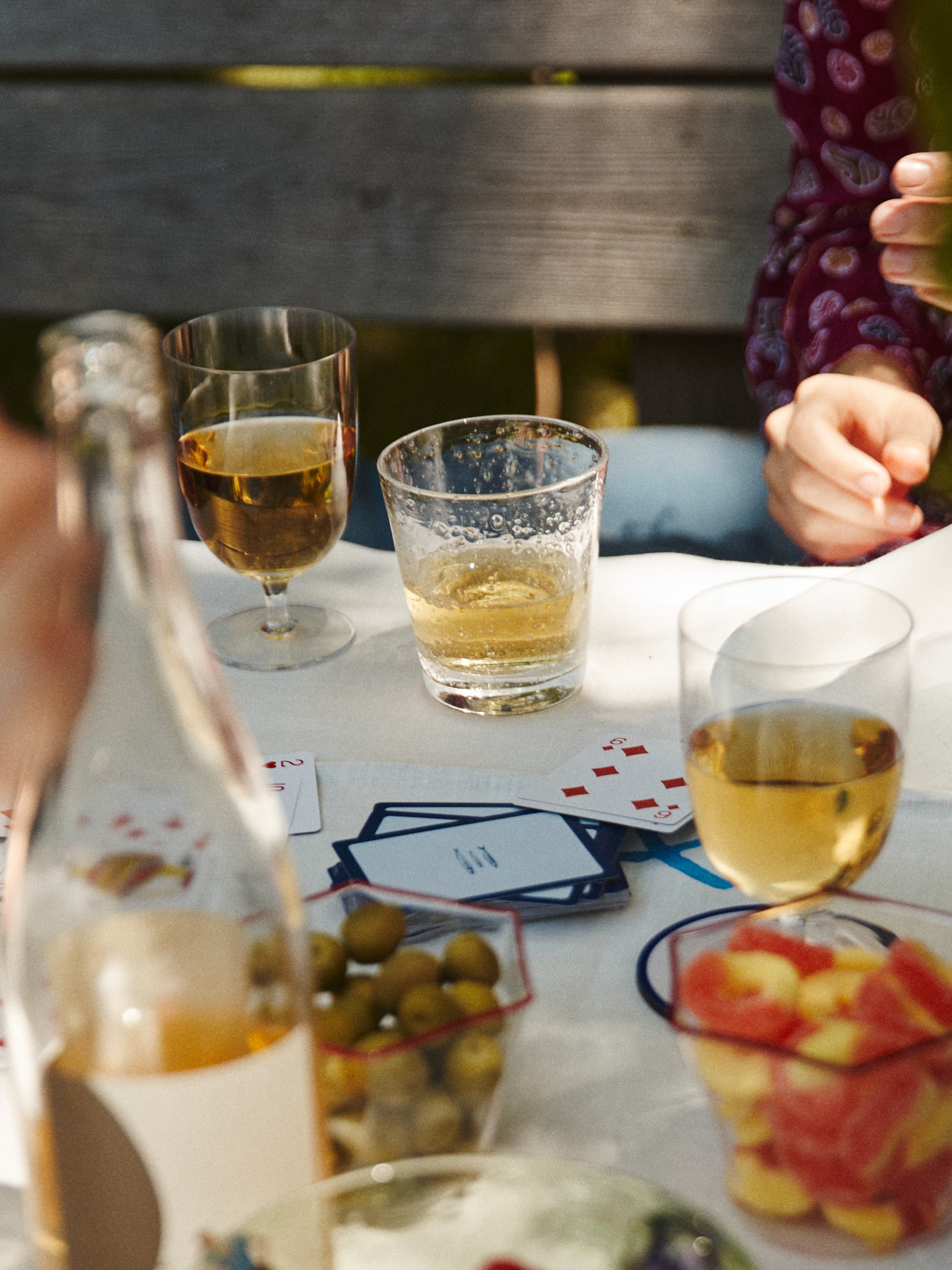 A table set with Lauren Water Glasses, a bottle, bowls of olives and fruit, scattered playing cards, drinks, and people sitting around.
