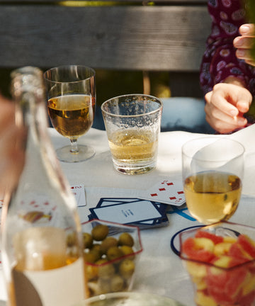 A table set with Lauren Water Glasses, a bottle, bowls of olives and fruit, scattered playing cards, drinks, and people sitting around.