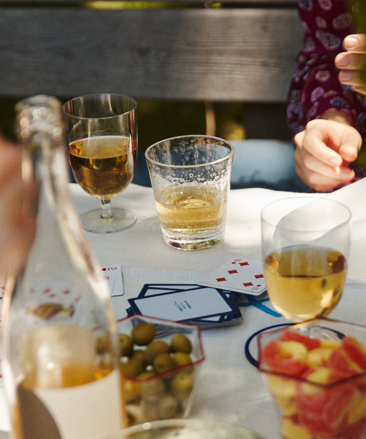 A table set with Lauren Water Glasses, a bottle, bowls of olives and fruit, scattered playing cards, drinks, and people sitting around.