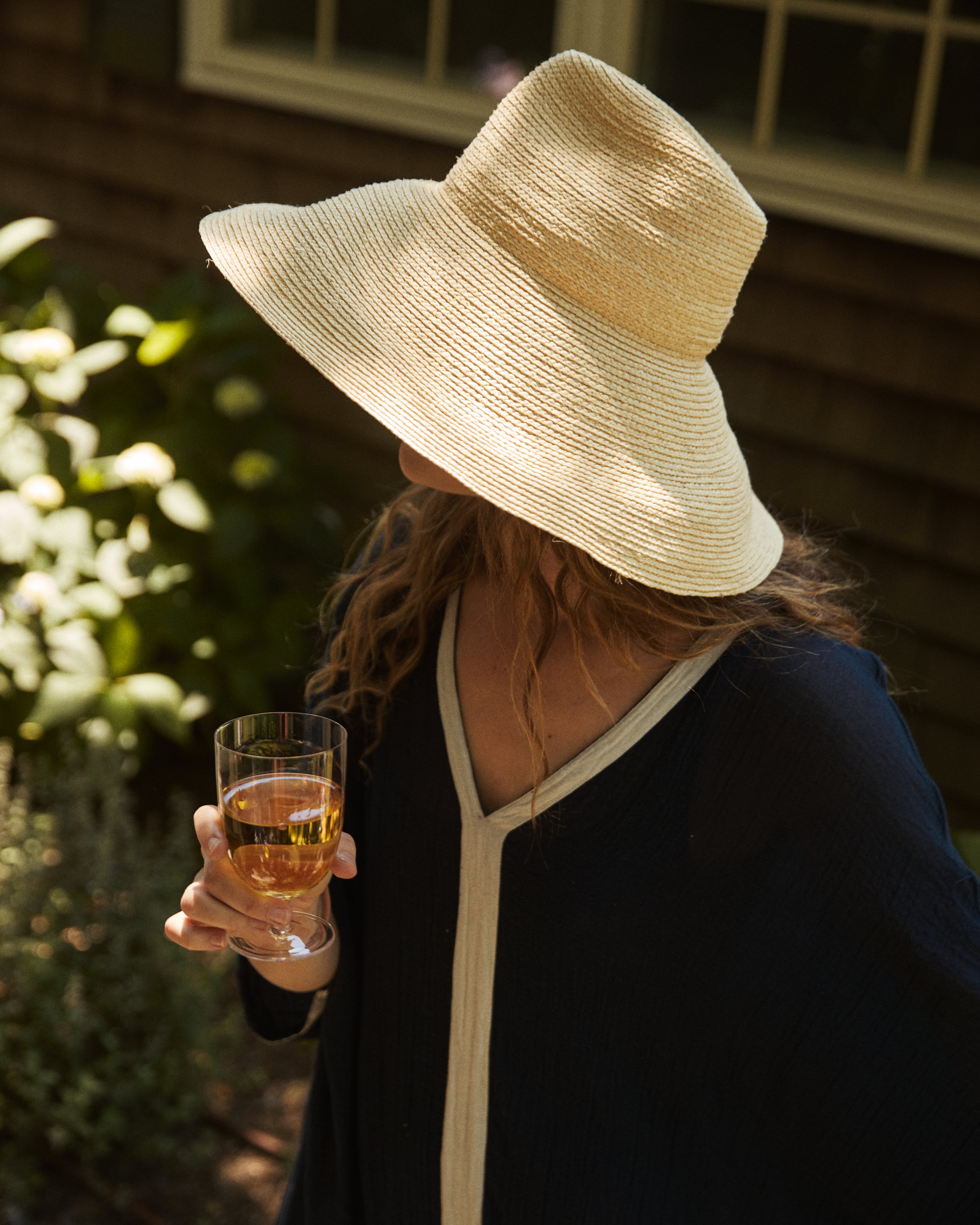 A person in dark clothing and a wide-brimmed straw hat enjoys the outdoors near a building, holding a light-colored drink in one of our Venezia Wine Glasses from the Set of 6.