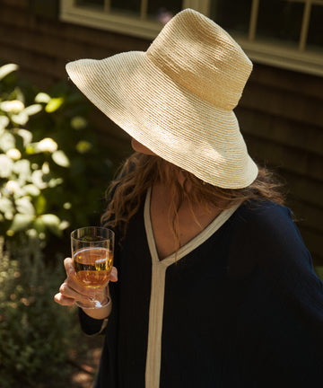 A person in dark clothing and a wide-brimmed straw hat enjoys the outdoors near a building, holding a light-colored drink in one of our Venezia Wine Glasses from the Set of 6.