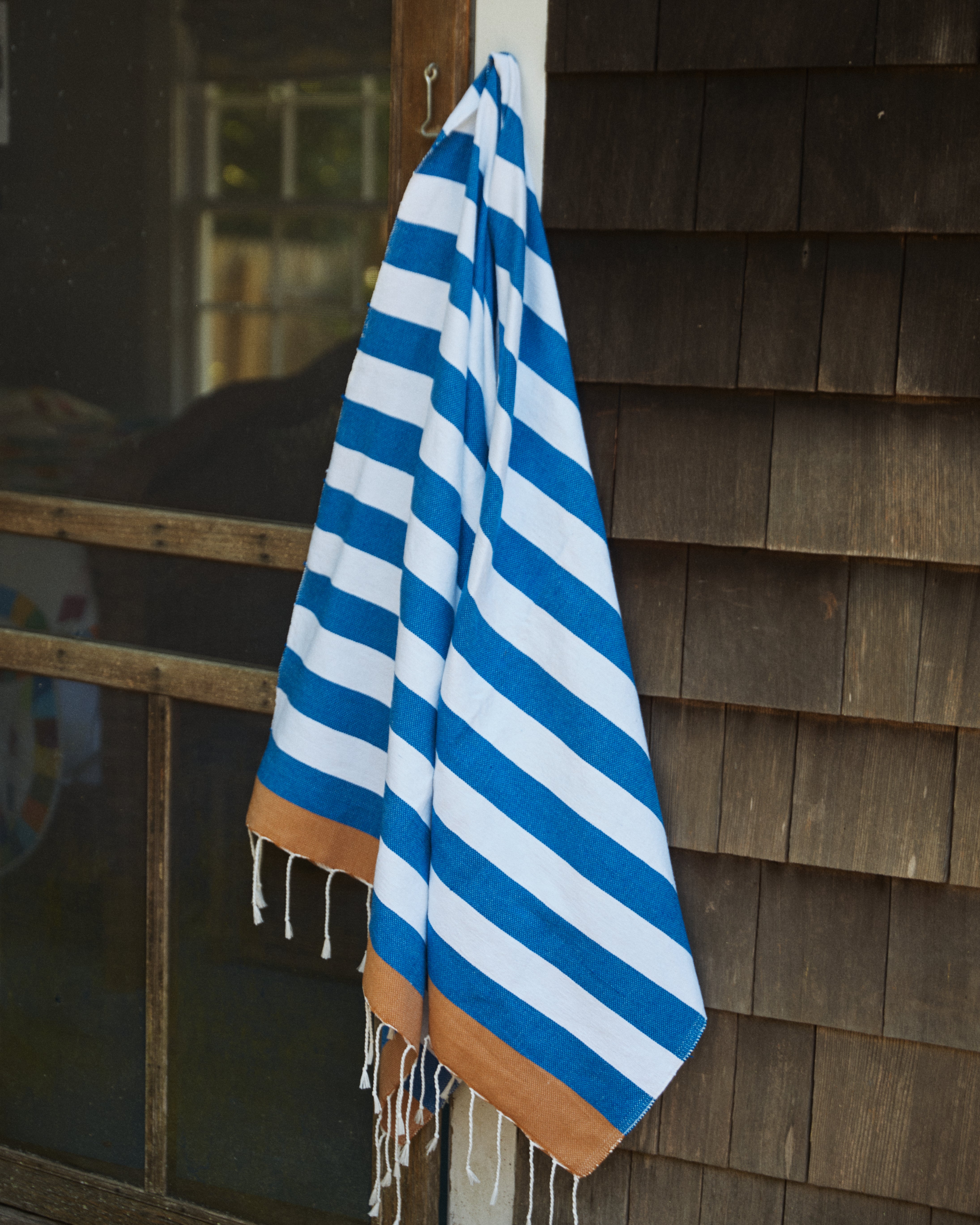The La Plage Towel, featuring blue and white stripes with brown ends, hangs on a hook outside next to a wooden shingle wall and screened door.