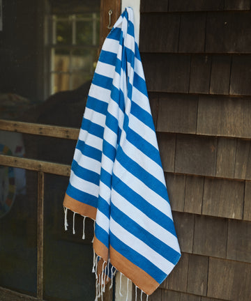 The La Plage Towel, featuring blue and white stripes with brown ends, hangs on a hook outside next to a wooden shingle wall and screened door.