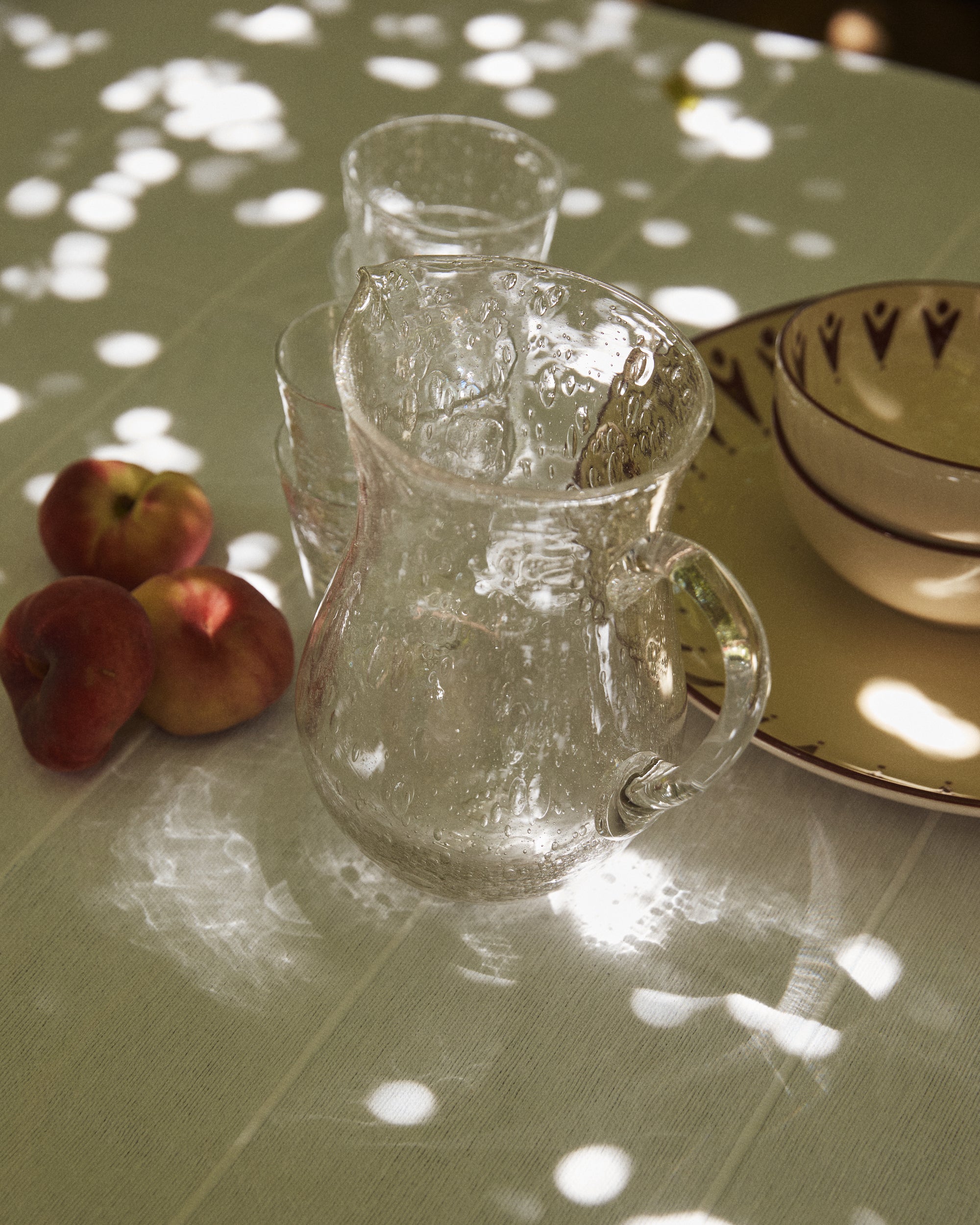 The Lauren Water Jug and two glasses rest on a white tablecloth beside three peaches and some dishes, with dappled sunlight casting patterns across the table.