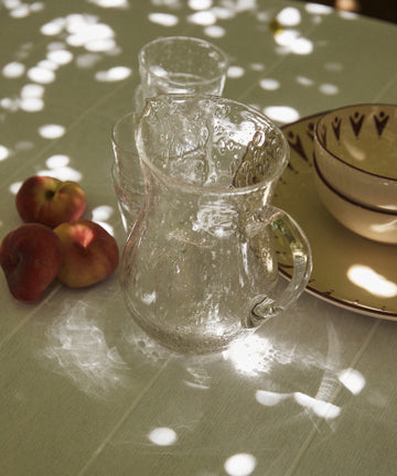 The Lauren Water Jug and two glasses rest on a white tablecloth beside three peaches and some dishes, with dappled sunlight casting patterns across the table.
